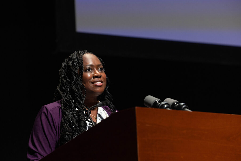 Woman in purple jacket, long braids speaks at podium