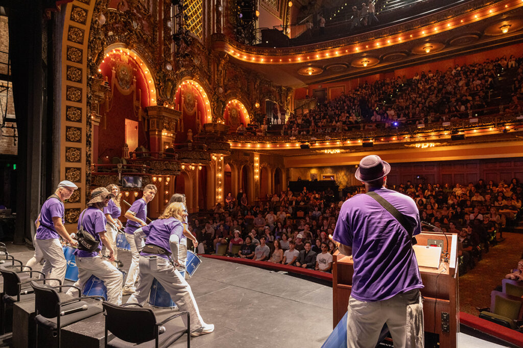 Drummers in purple t-shirts play on stage as audience in ornate theater looks on