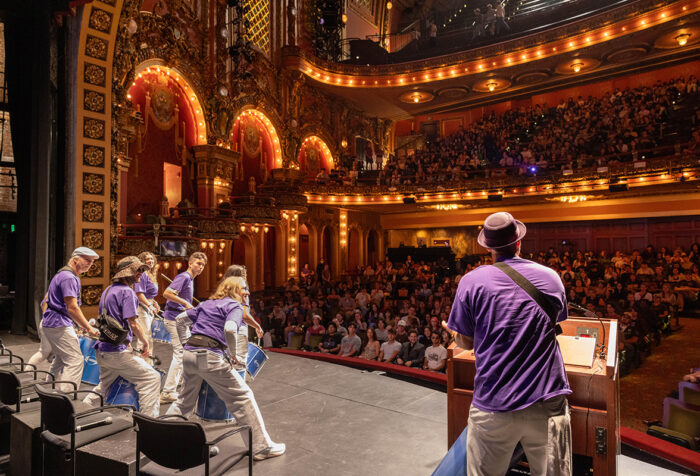 Drummers in purple t-shirts play on stage as audience in ornate theater looks on