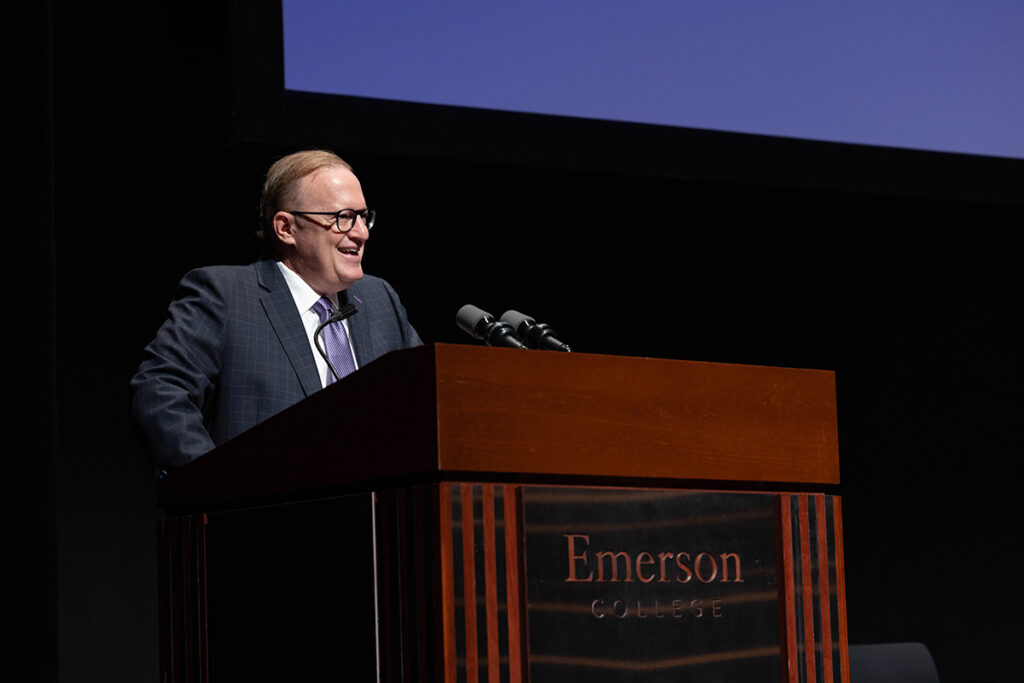 Man in blue suit, purple tie, glasses speaks at podium