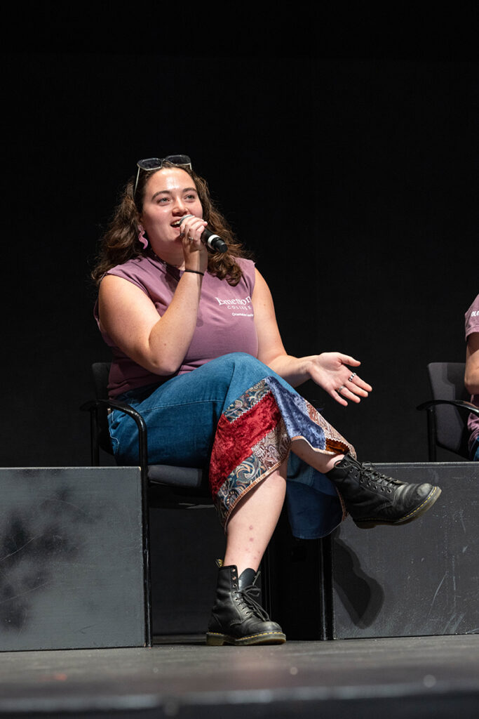 Student in purple Emerson T-shirt, denim skirt, black boots speaks into microphone on stage