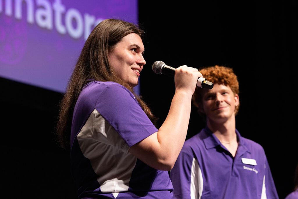 Student in purple shirt, long brown hair speaks into microphone as student in purple shirt, curly red hair looks on