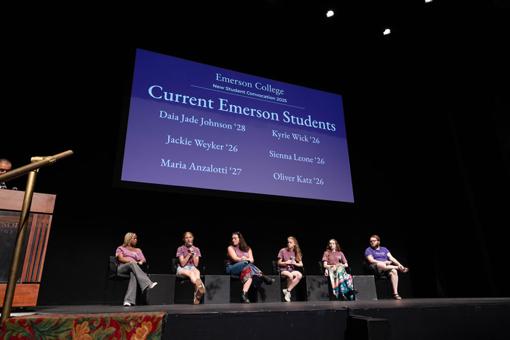 Six students in purple Emerson T-shirts sit on stage with microphones, their names projected on a screen behind them.