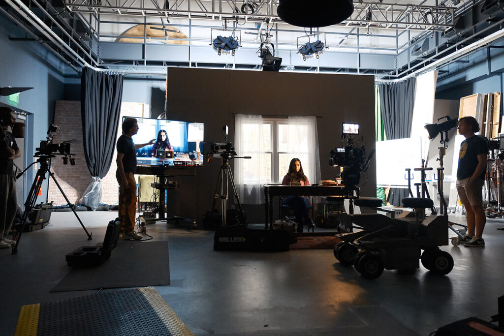 woman sits at desk on film set as man points to her image on a screen and another looks on