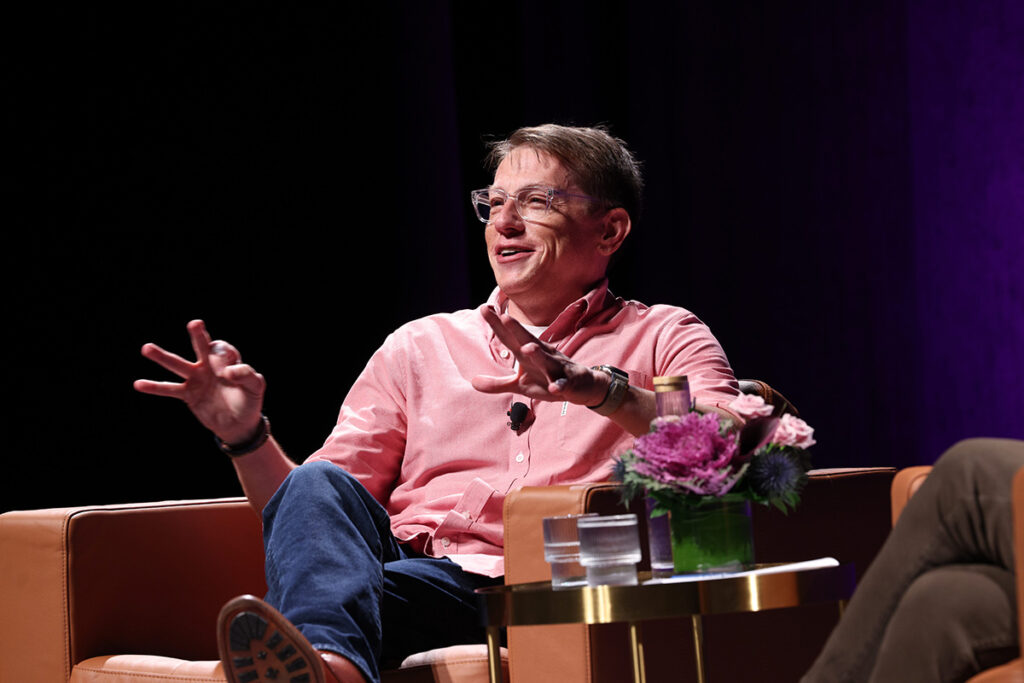man in pink shirt, jeans gestures while sitting in a chair on stage