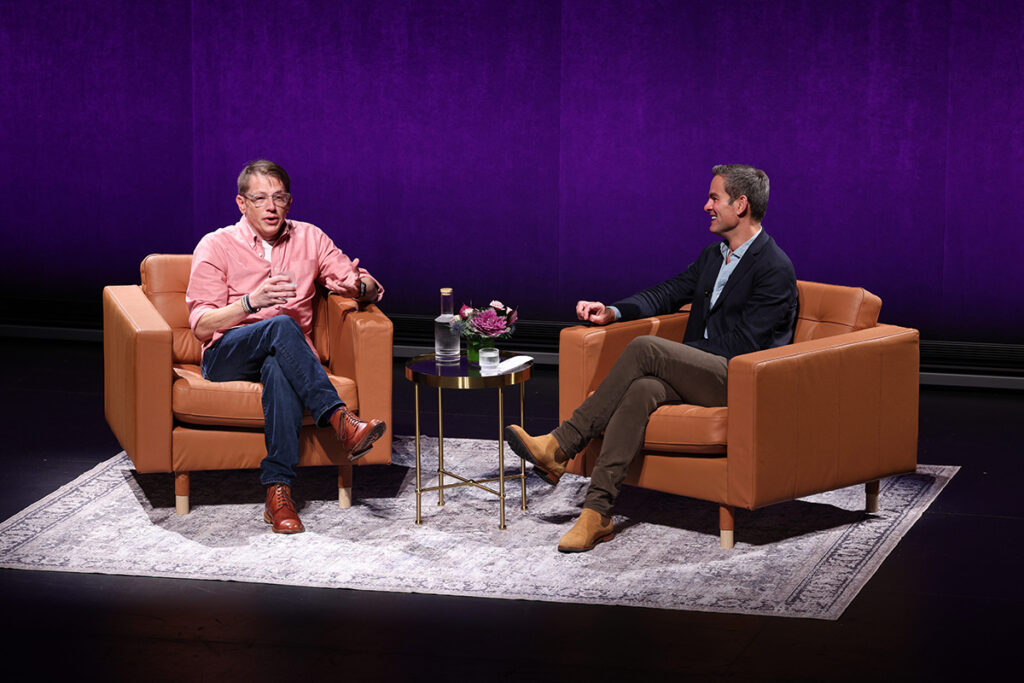 Two men sit in brown leather chairs on a stage backed in purple