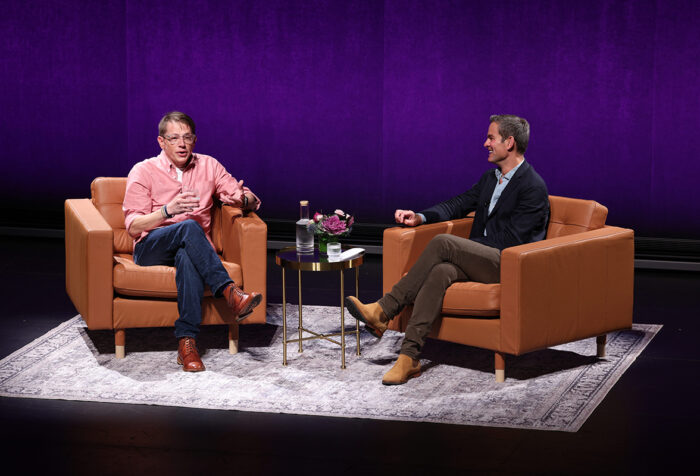 Two men sit in brown leather chairs on a stage backed in purple