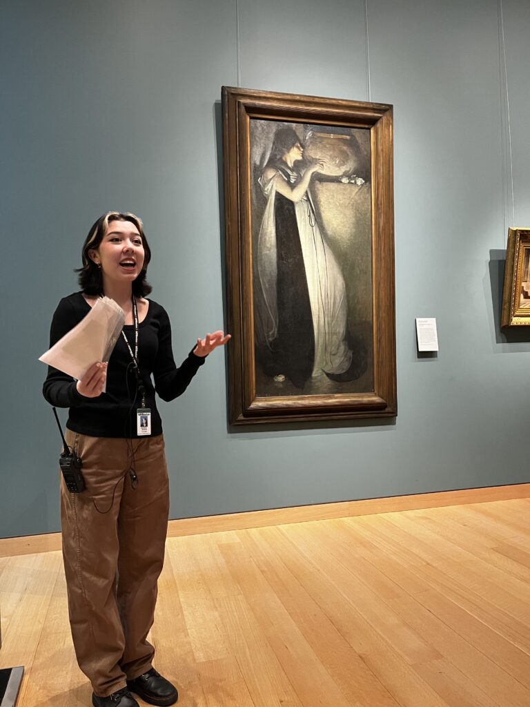 Madalyn Jimiera provides a presentation at the Museum of Fine Arts with a painting behind her on the wall.