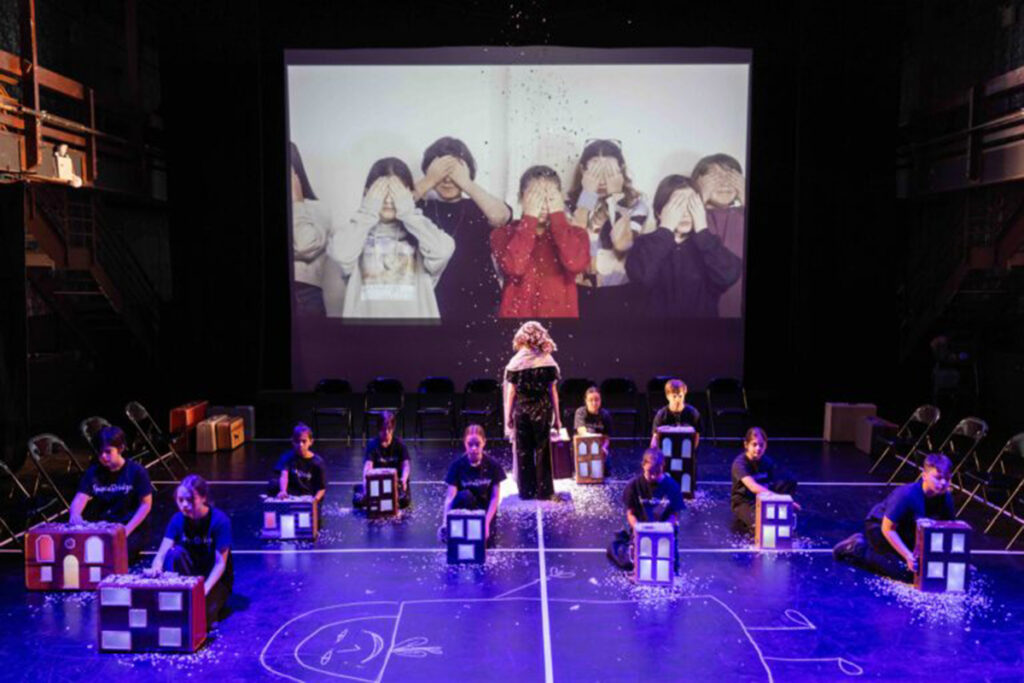 Children kneel on stage behind cardboard models of houses, children on screen behind them cover their eyes