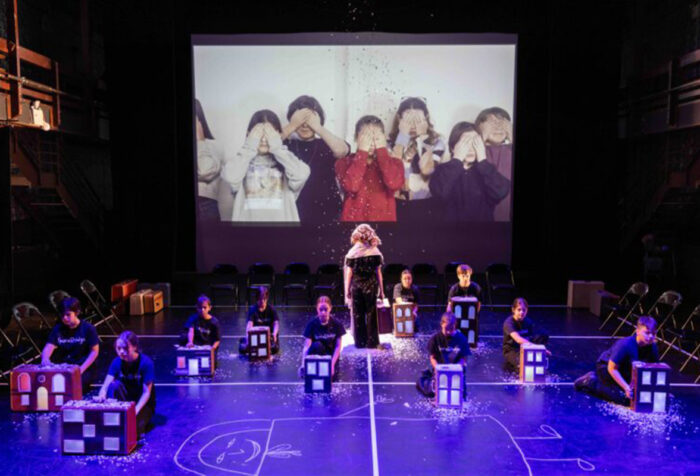 Children kneel on stage behind cardboard models of houses, children on screen behind them cover their eyes