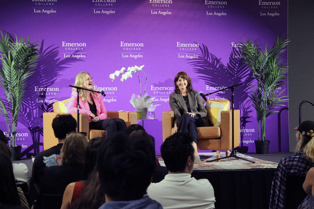 two women sit behind microphones in chairs as audience looks on