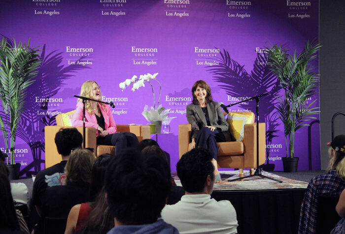 two women sit behind microphones in chairs as audience looks on