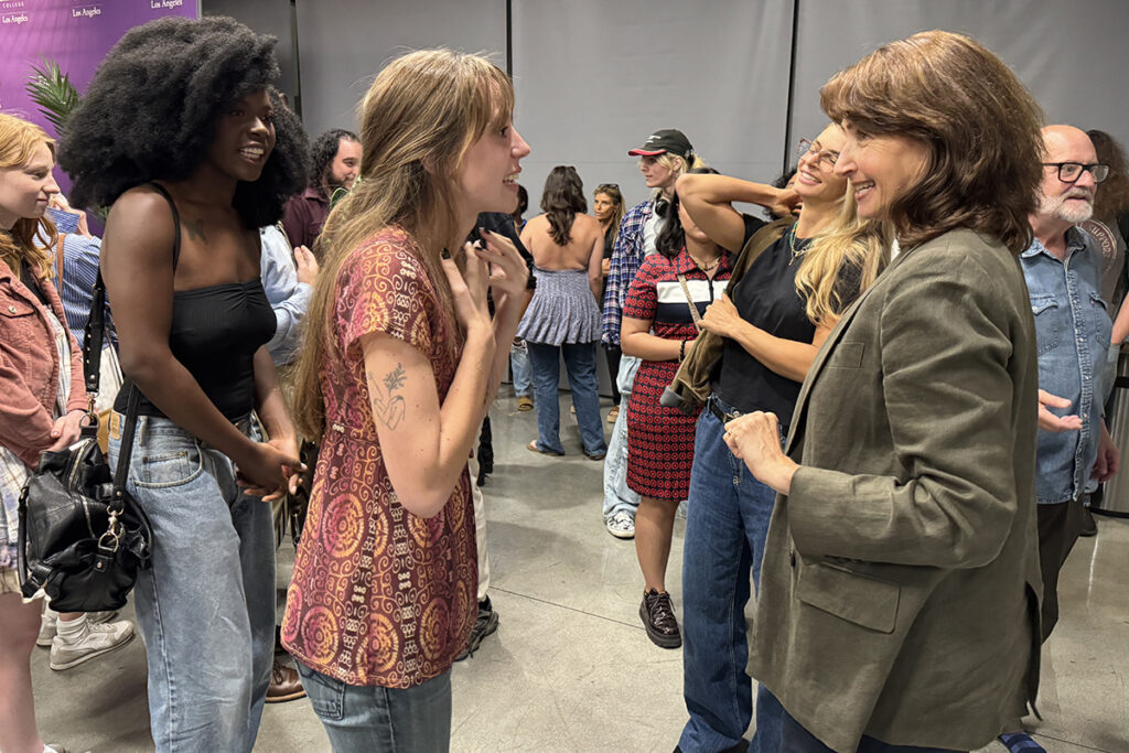 Student with long hair in pink shirt and jeans gestures as she talks to woman wearing green blazer