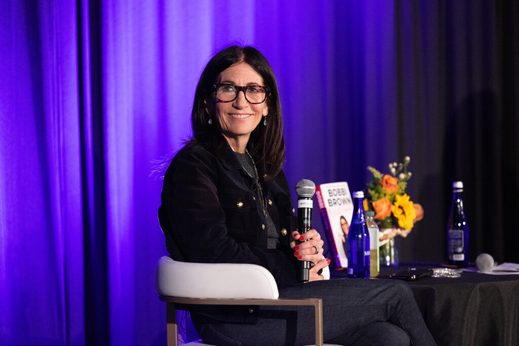 Woman in black top holding mic turns at angle in chair and smiles