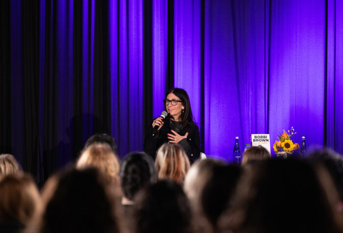 Woman in black top speaks into mic on stage in front of purple curtain. Audience heads in foreground