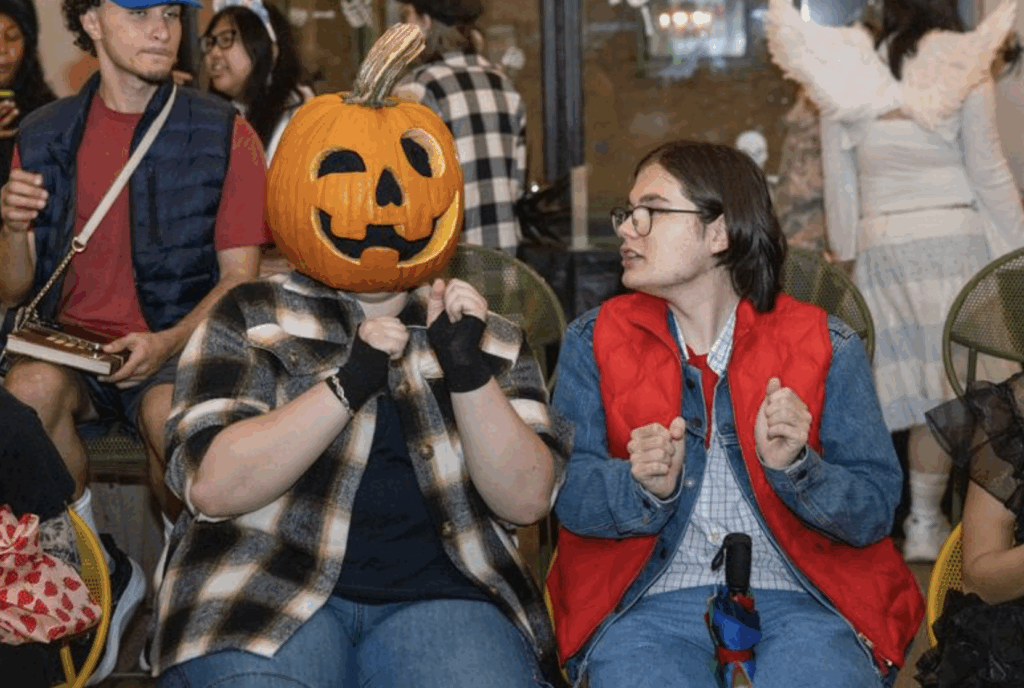 A person wearing a pumpkin on their head sits next to someone in a red vest