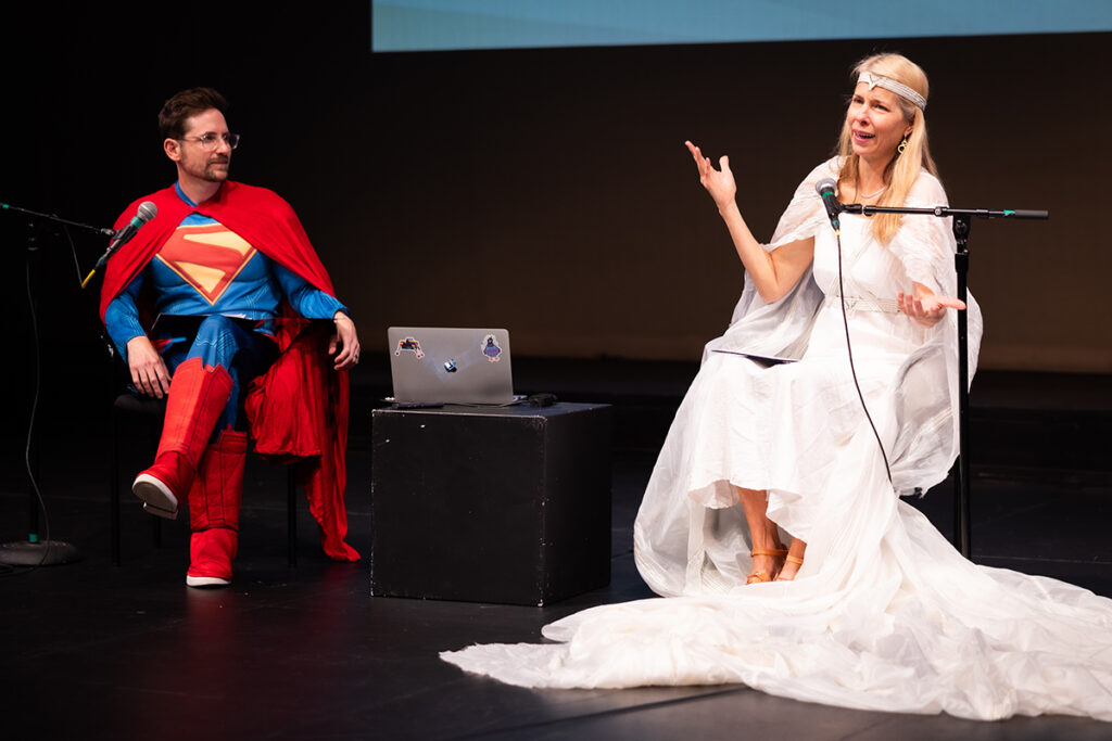 man in superman costume and woman in flowing white robes chat on stage