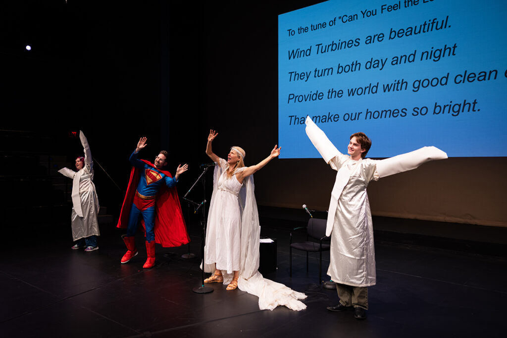 Man dressed as superman, woman dressed as Cassandra, two students dressed as windmills raise arms on stage