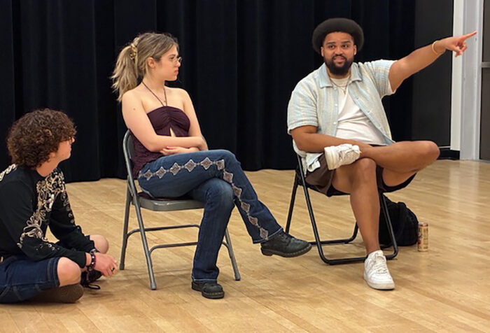 Kai Tshikosi points his hand up while sitting in a chair and two students listen to him