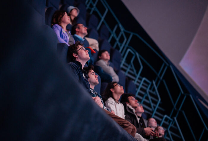 People in steeply pitched theater seats look up at screen