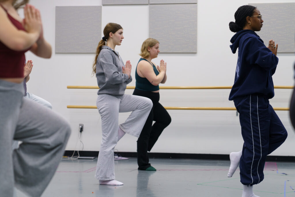 Students stand with hands to their chest in prayer form while standing on one leg