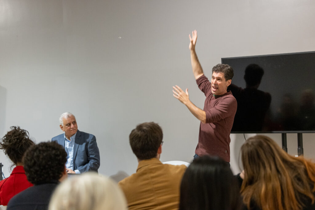 Max Brooks speaks to a class with his hands raised in the air