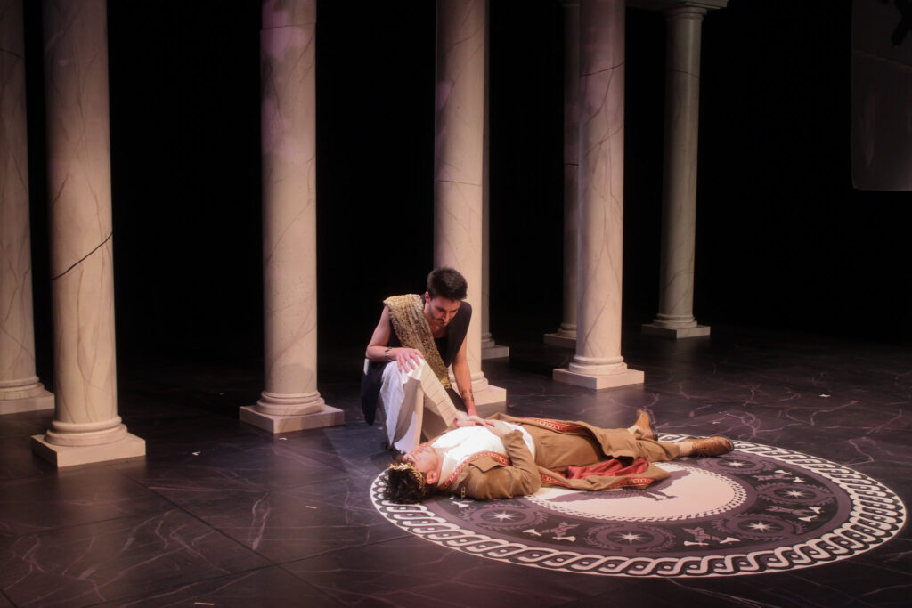 Two actors on stage during Julius Caesar. One of them lays on the floor, while the other kneels down.
