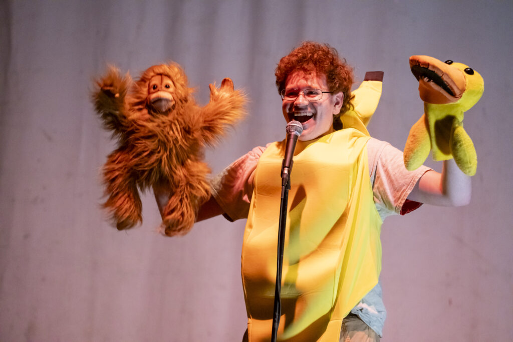 Ricky Downes performs stand up dressed as a banana while holding a puppet duck and a puppet orangutan. 