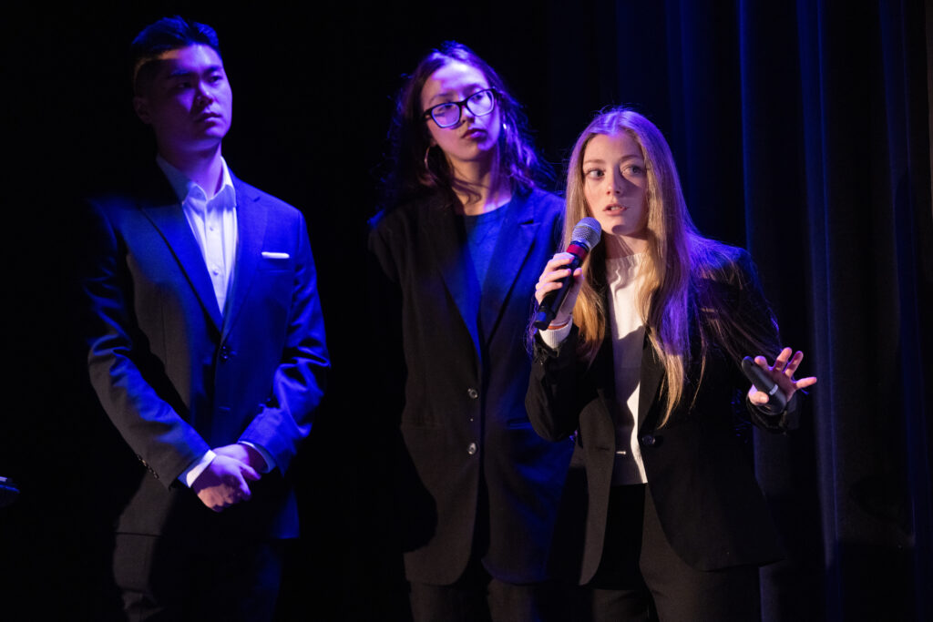 One person talks while holding a microphone and two people look on behind the person talking