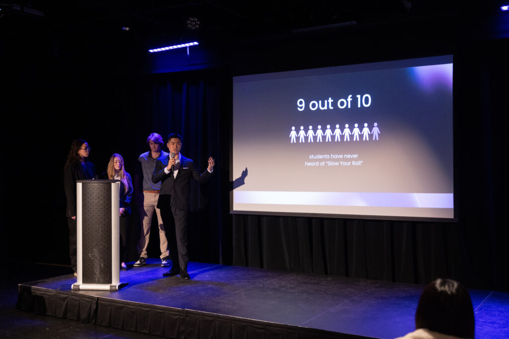 Four students on stage with one of them speaking while holding a microphone and there's a projection screen