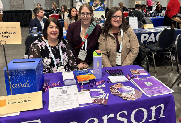 Three women stand behind table festooned with Emerson branded handouts at convention center