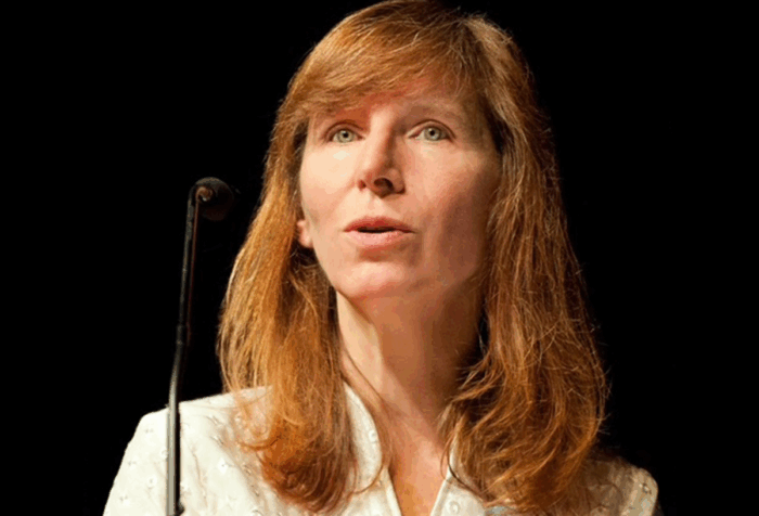 A person with red hair, wearing a white shirt, is speaking at a podium.