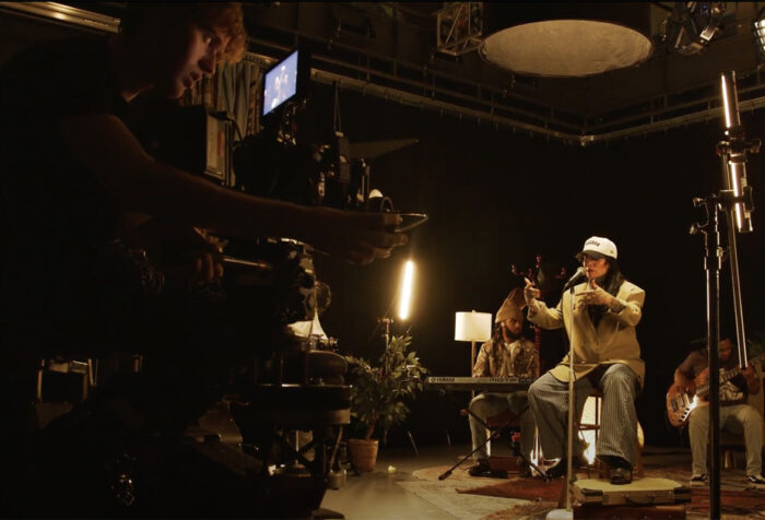 A cameraman sits on a dolly operating a camera while musician Lisa Bello and her band perform live on a set