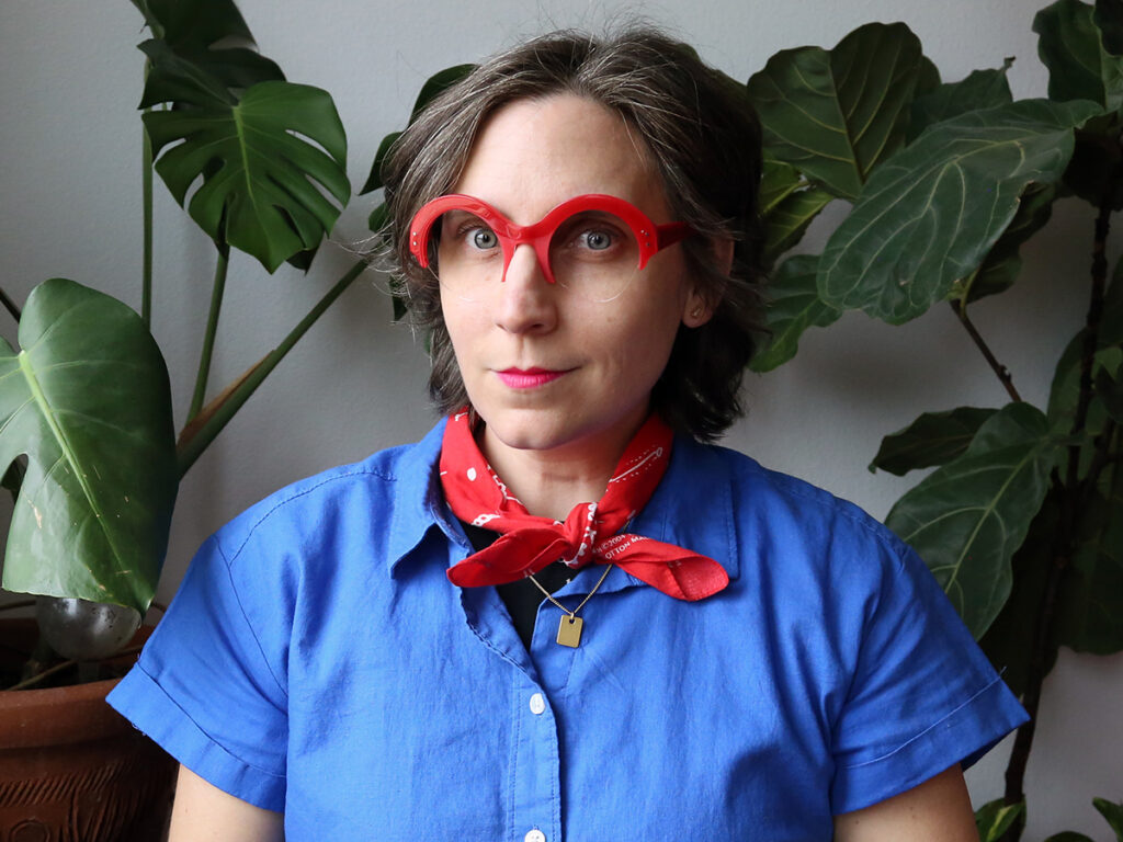 Liss LaFleur, wearing a blue button-down shirt, red neckerchief, and round red-framed glasses