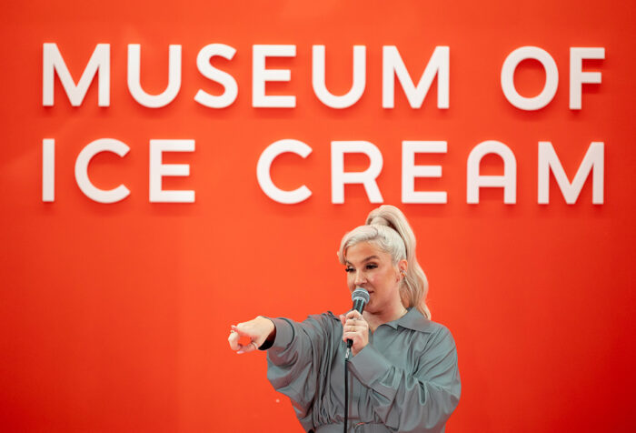 Joelle James points while singing into a microphone with Museum of Ice Cream on the wall behind her