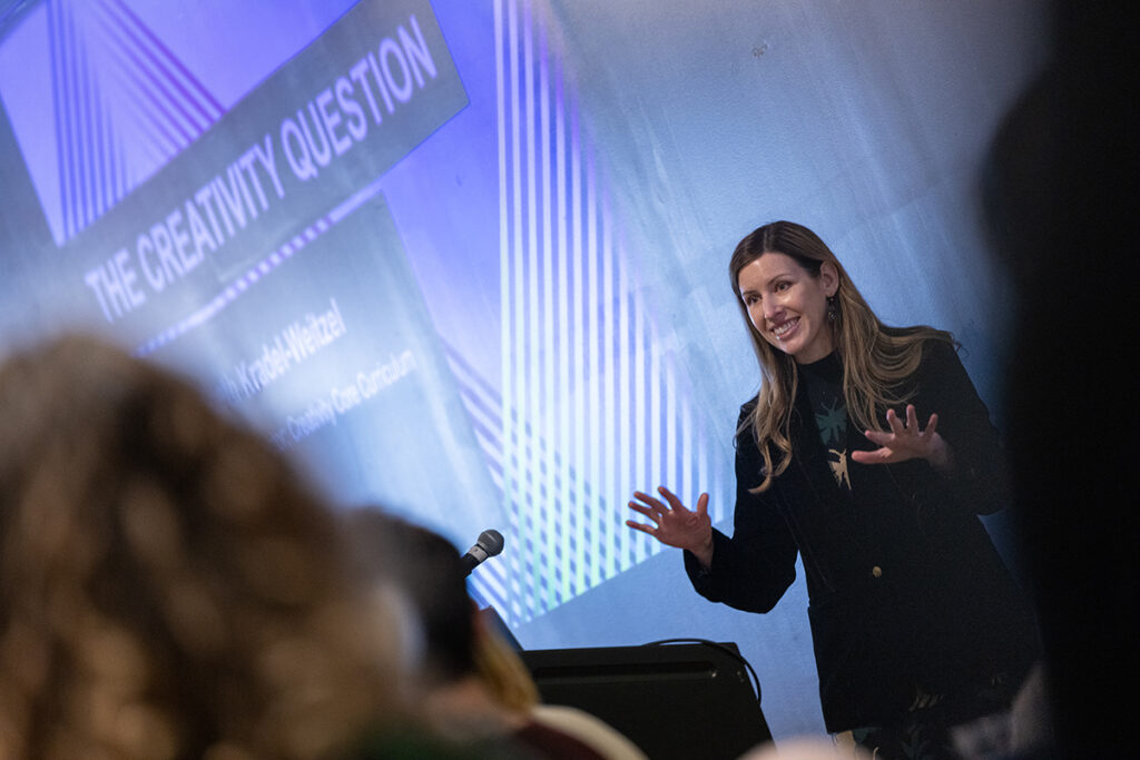 Woman gestures in front of a screen reading "The Creativity Question"