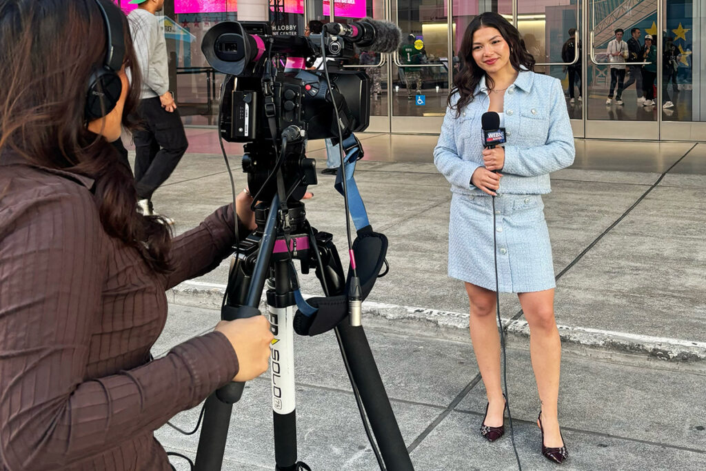 Woman in pale blue skirt suit does standup in front of glass doors as woman in brown shirt films