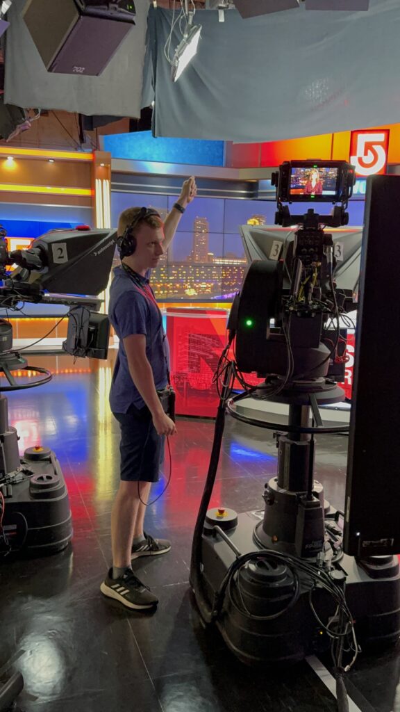 Aidan Vahey raises his hand next to a TV studio camera 