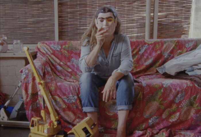 Image shows a woman sitting on a couch, holding up a mask in front of her face.