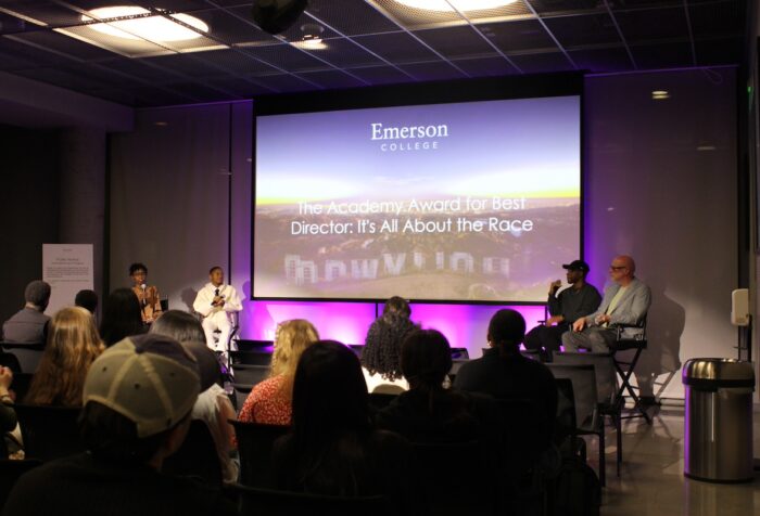 Panelists sit at the front of the room, with a large screen behind them while audience members, facing the panelists, listen