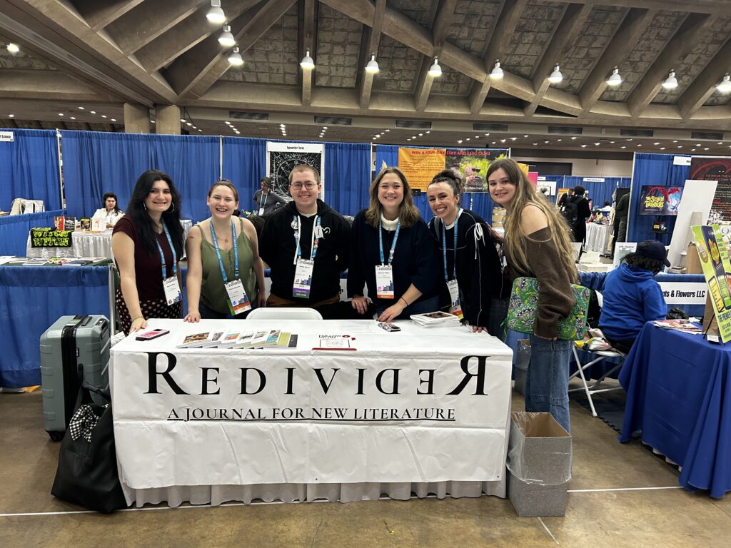 Six staff members of Redivider stand for a photo behind the magazine's table