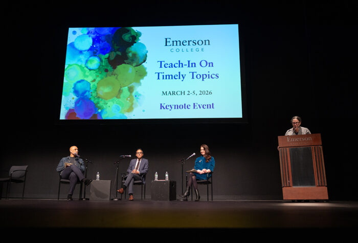 Three people sit in chairs while one person stands at a lectern with a big projection screen behind them