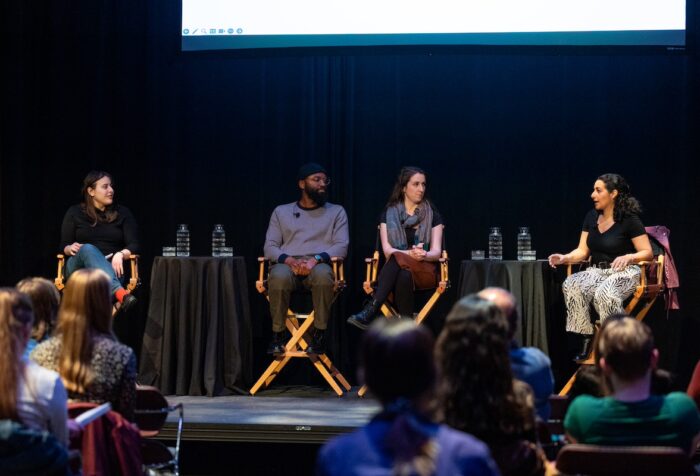 Four panelists seated in directors' chairs on a stage