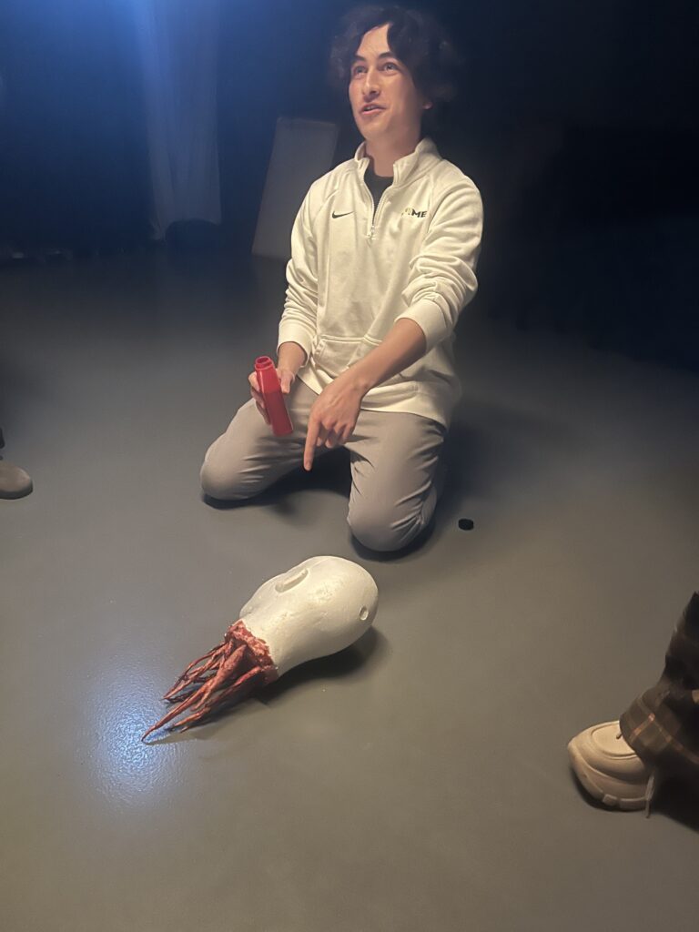 Max Gellen kneels on the ground with a mannequin head on the floor with entrails coming out of it