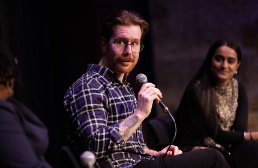 Nicholas Medvescek speaks while holding a microphone and sitting