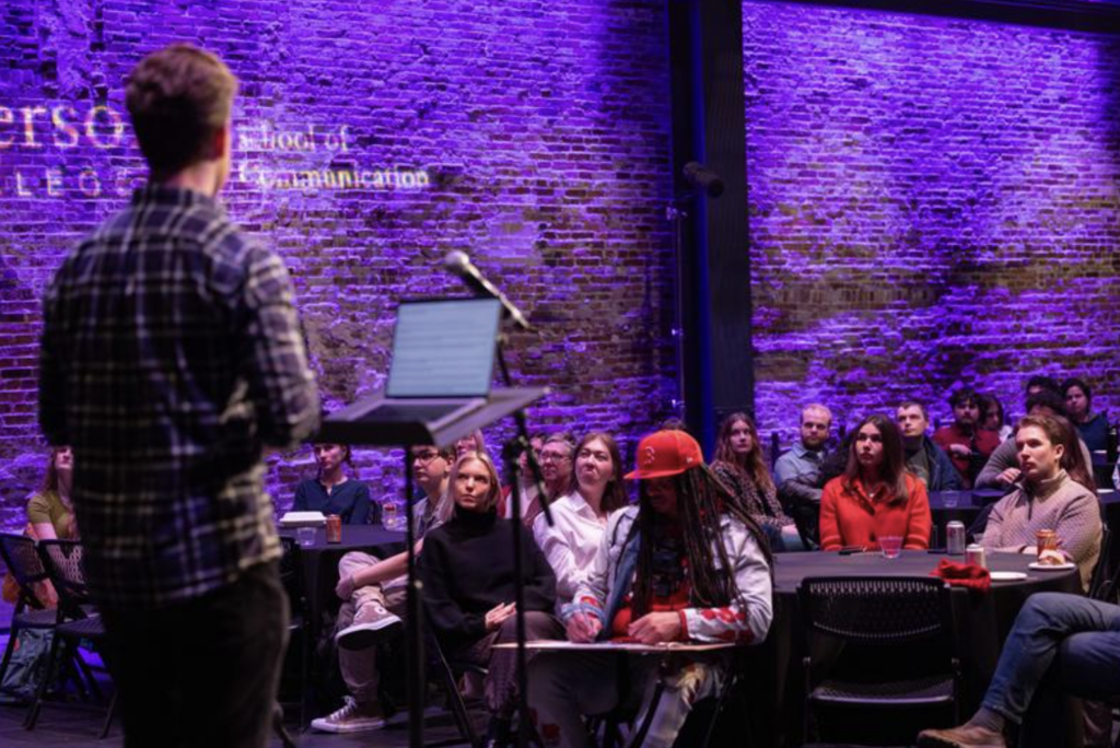 The view is of the audience looking at a man standing in front of a lectern from behind the man