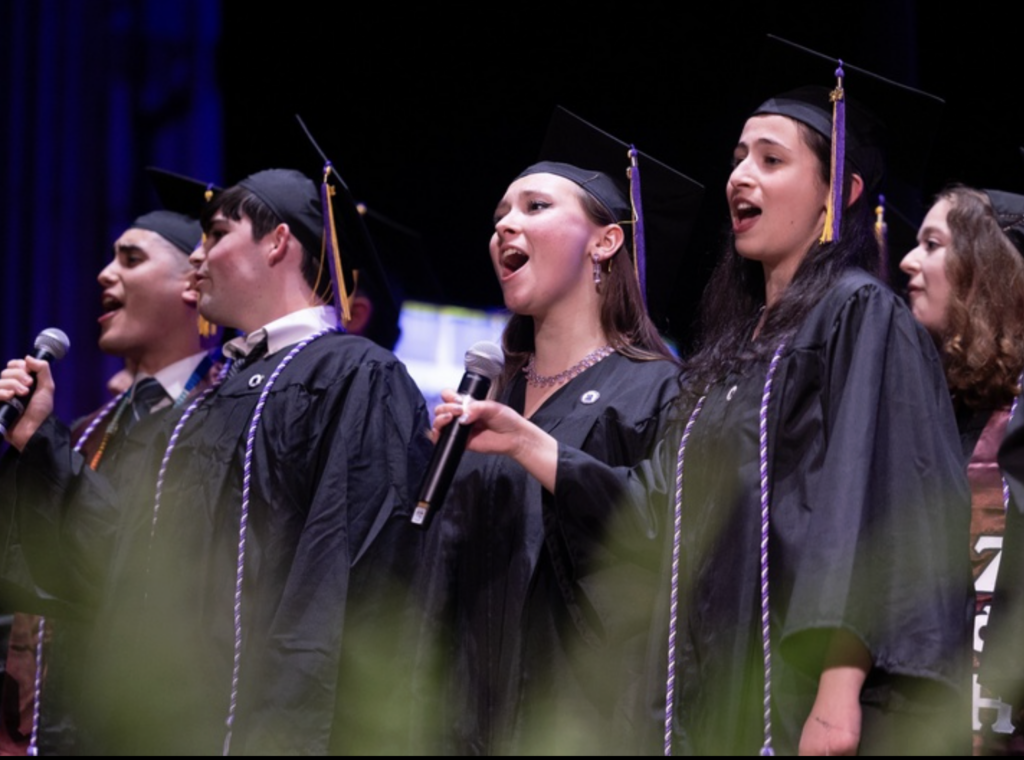 Students sing on stage while wearing cap and gowns