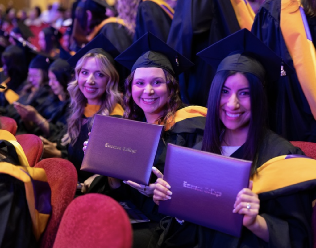 Graduates sit while holding their diplomas and smile