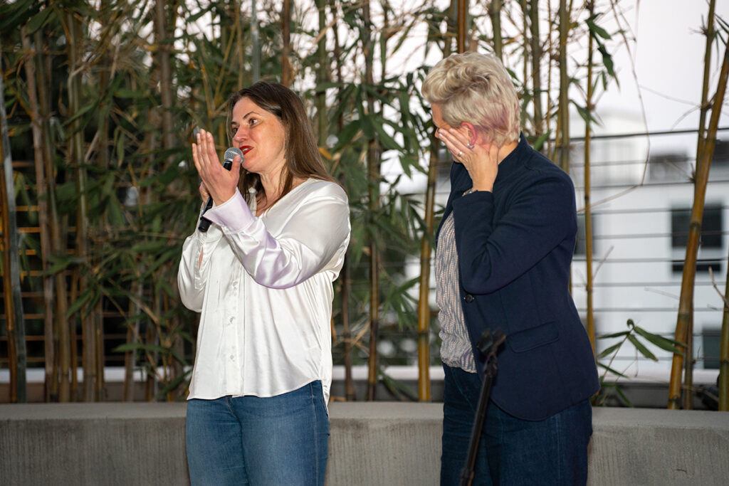 Woman speaks into mic in front of bamboo as another woman looks on