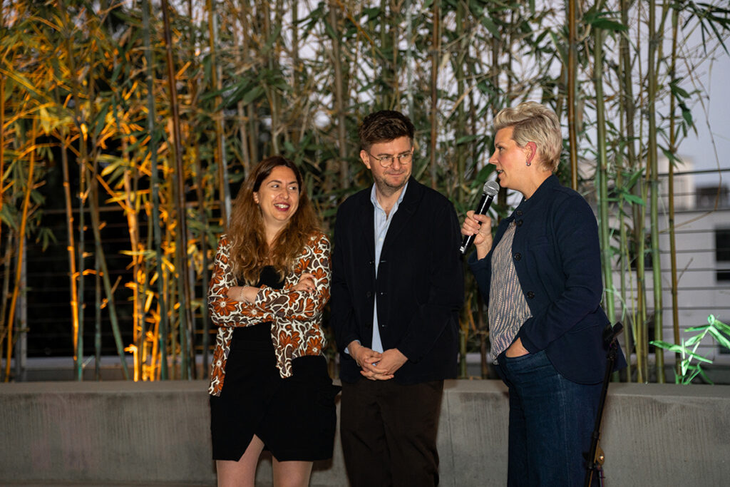 Woman with short blonde hair speaks into mic in front of bamboo plants as woman, man stand next to her.